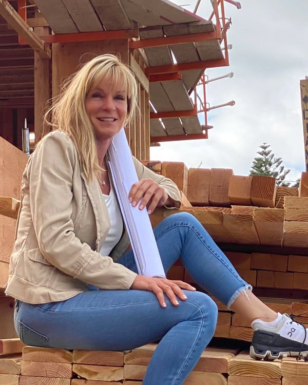 Dawn, woman general contractor, sits in tan blazer on a lumber pile with blueprints in her hand, smiling, ready to help coach